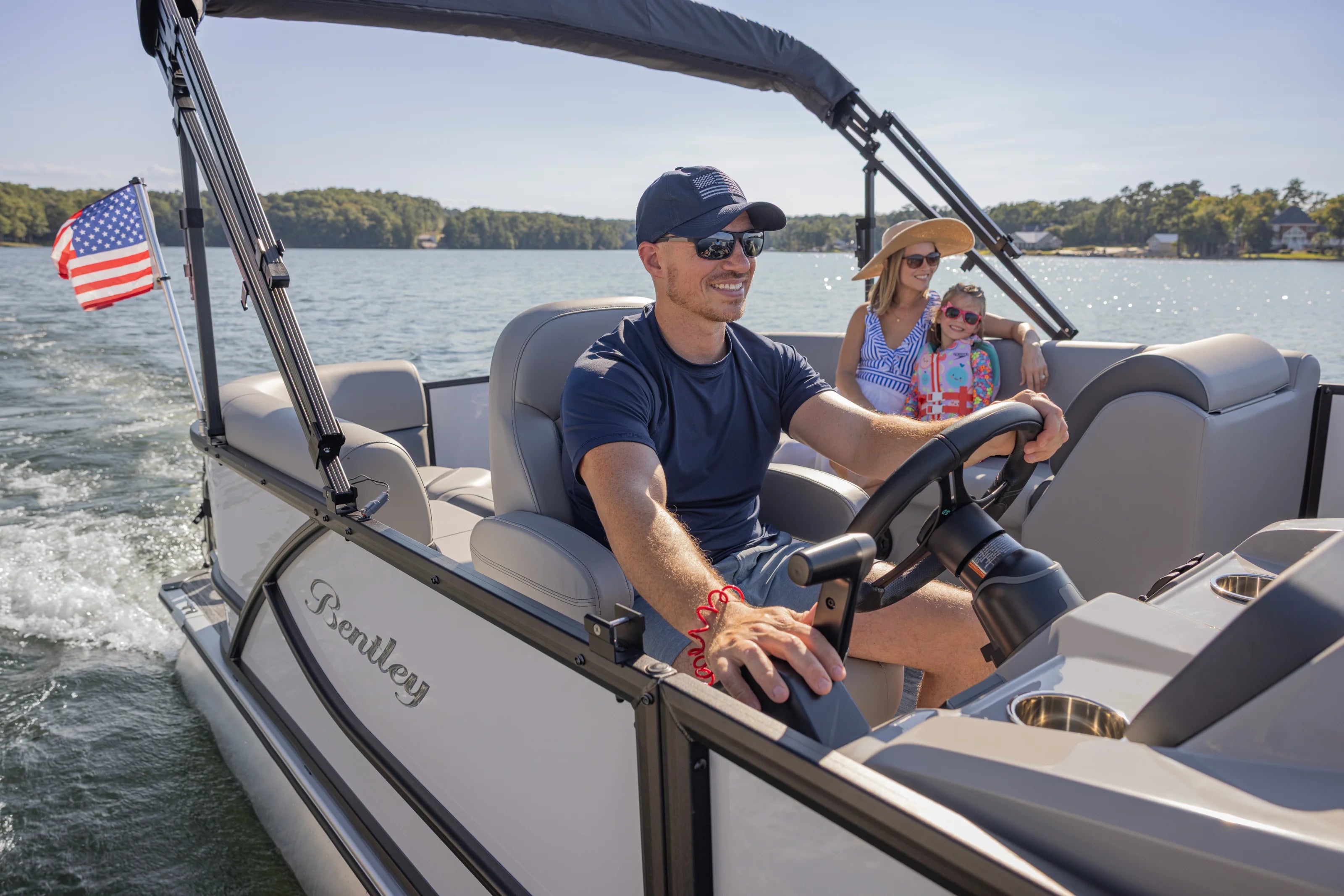 Man driving a Bentley boat on a lake with family in the background