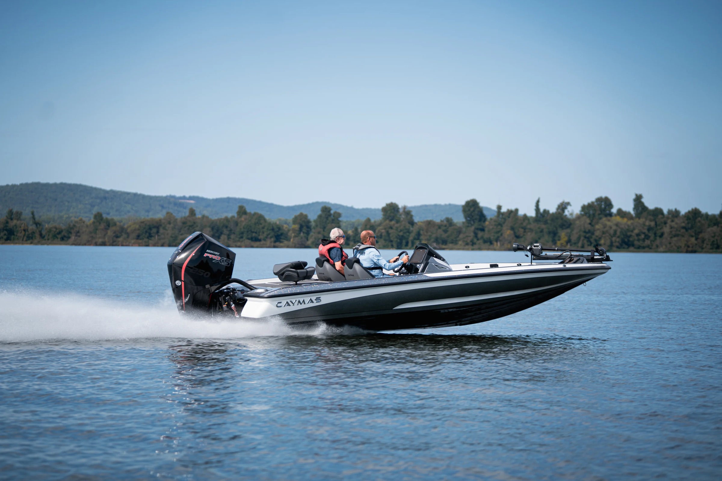 Two people on a white and black Caymas fishing boat cruising on a lake, surrounded by trees and mountains.