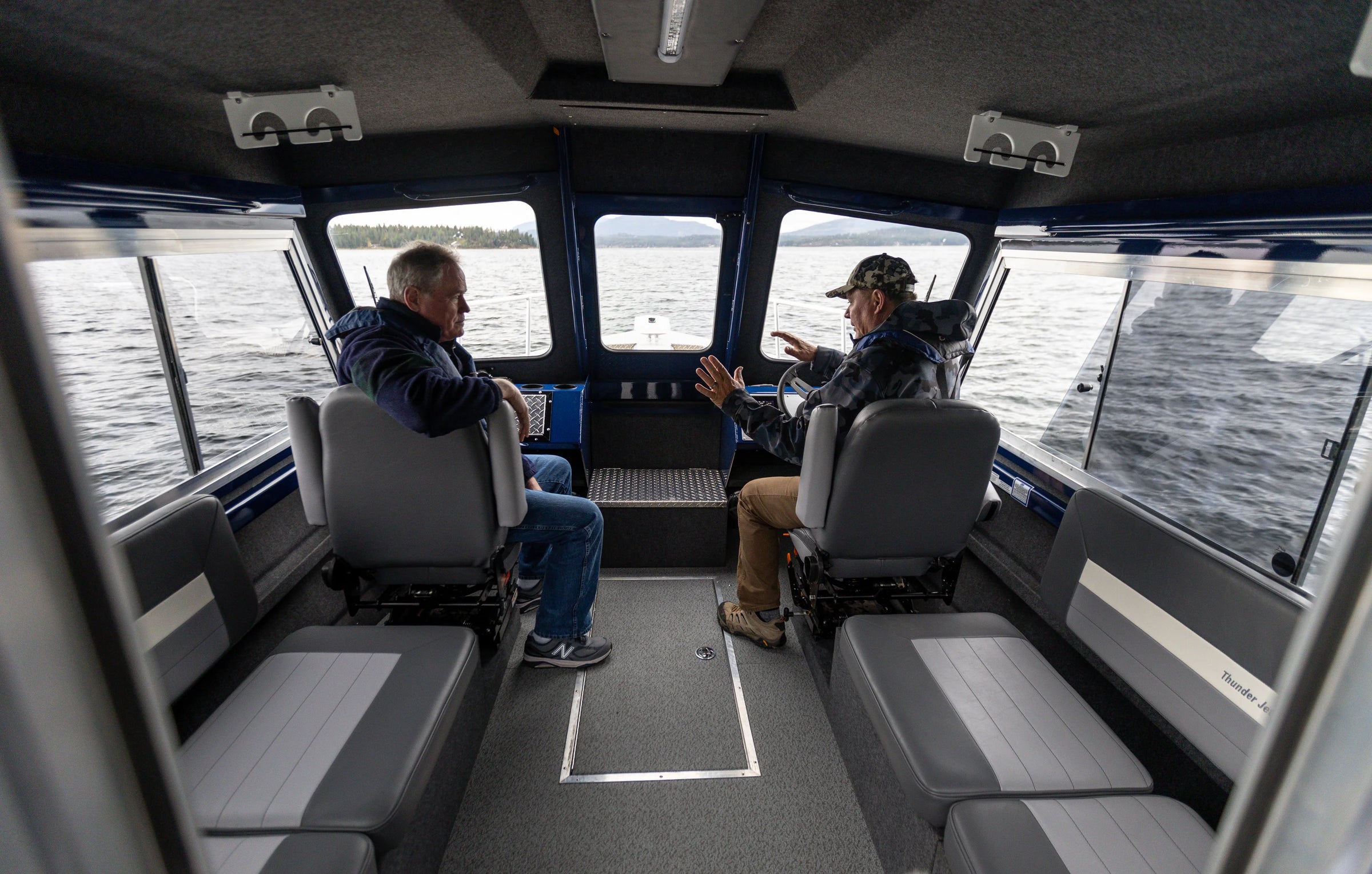 Two people inside a boat cabin with a view of water through the windows.