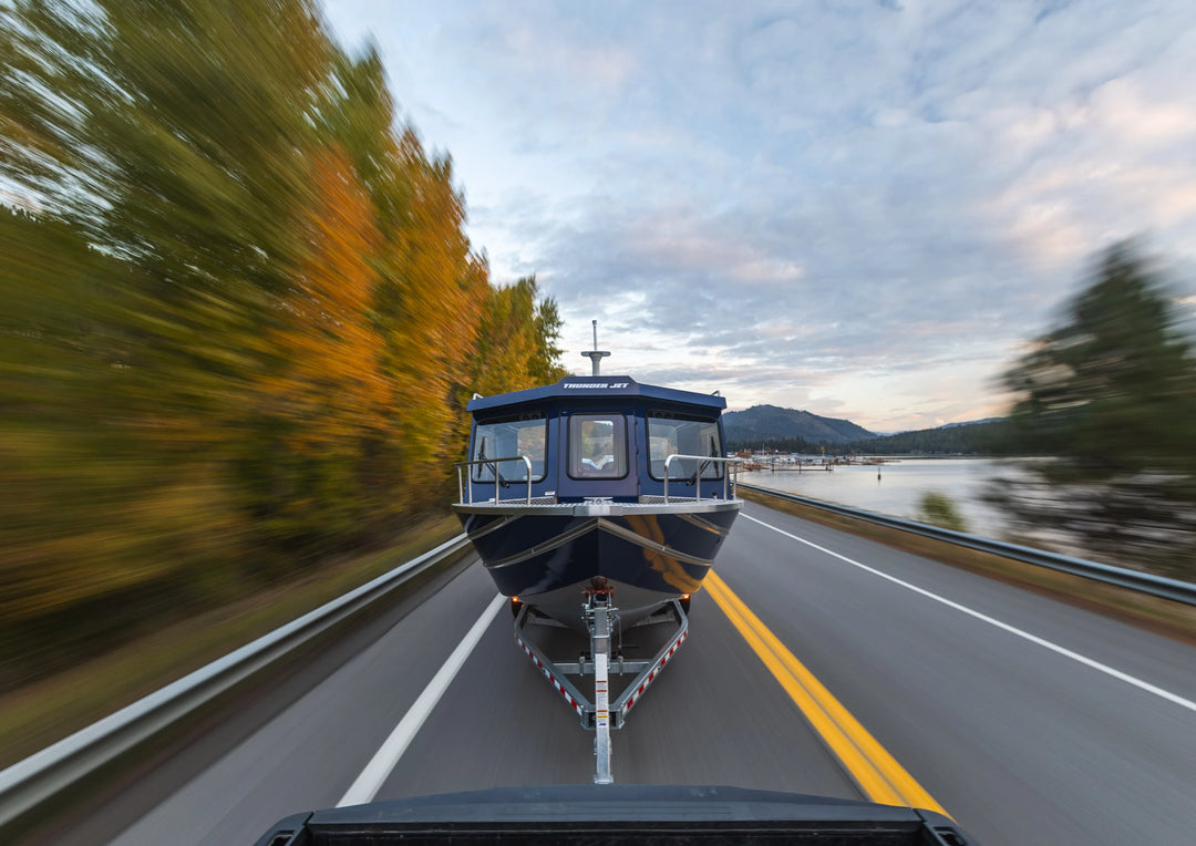 Boat on a trailer being towed on a road with trees and water in the background